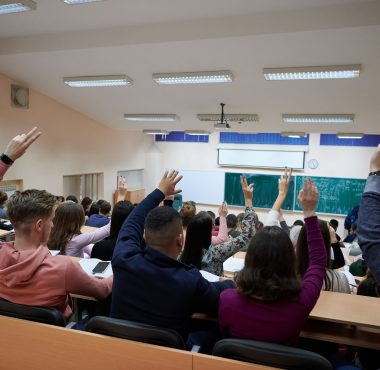 a large group of students raises their hand to answer a question asked by the professor